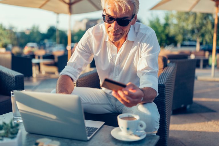 A senior man books a flight on his laptop at an outdoor cafe