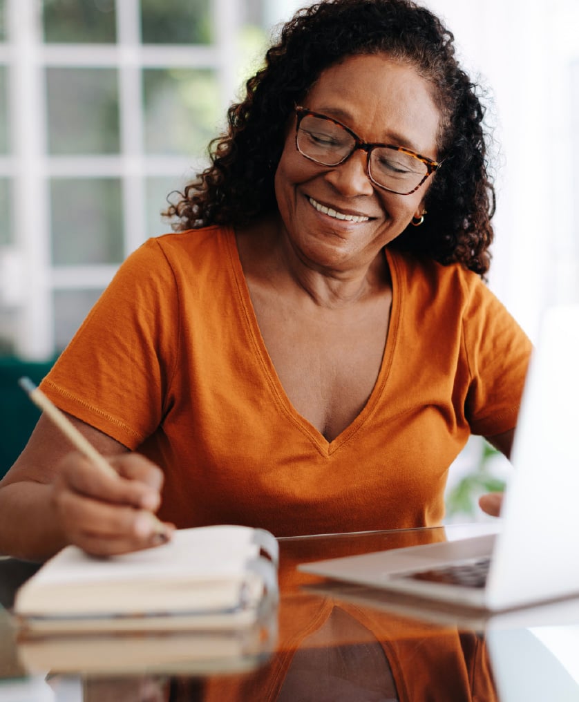 a photograph of a happy senior african american woman wearing an orange shirt and glasses, jotting notes in a notebook and using a laptop